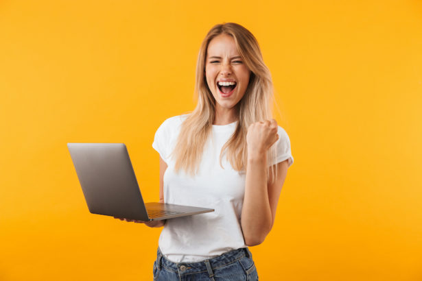 Portrait of an excited young blonde girl holding laptop computer and celebrating success isolated over yellow background